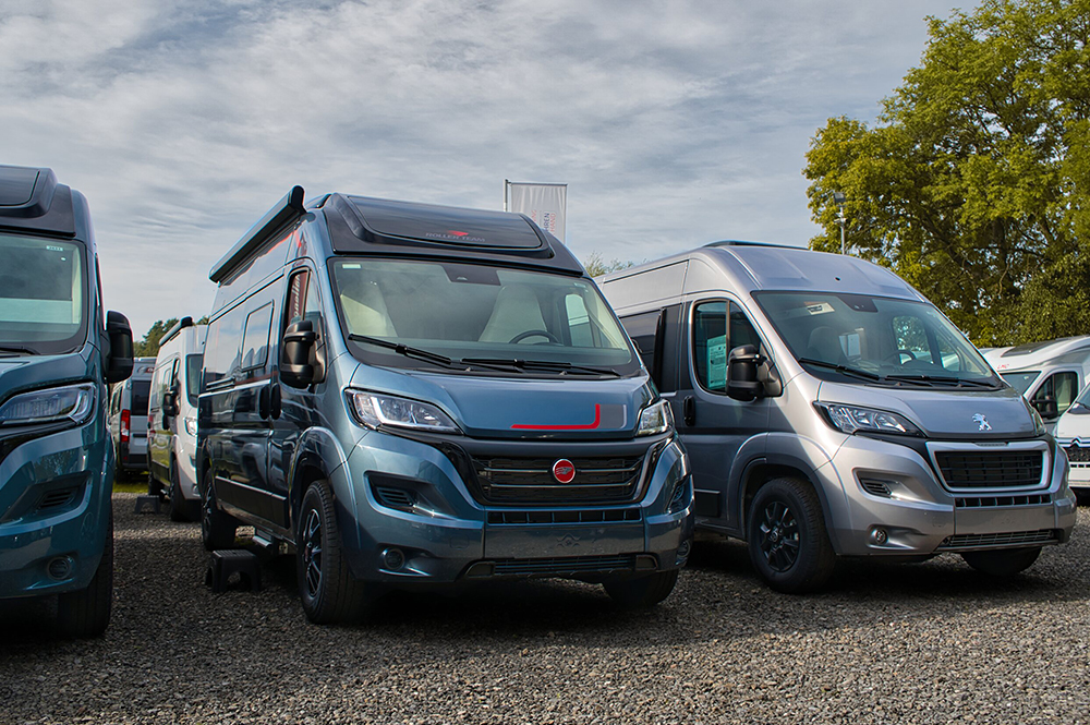 Mehrere Campervans nebeneinander auf einem Kiesparkplatz.